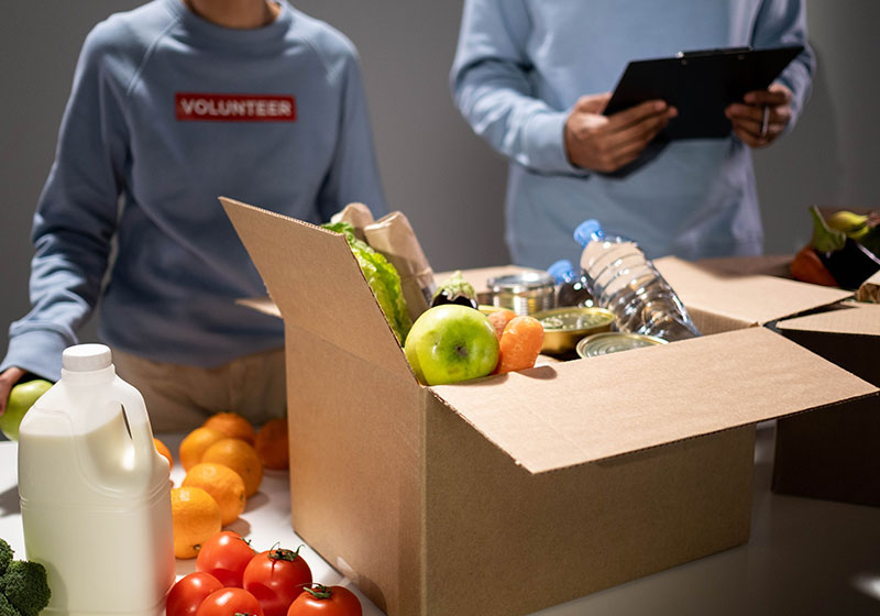 caixa de papelão com frutas e legumes e duas pessoas ao fundo